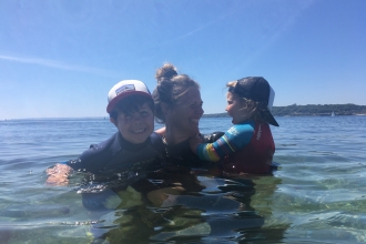 Marine Team Officer Abby Crosby bobs in the water with her two young children, surrounded by clear,calm blue waters and a bright blue sky behind. They are all laughing happily and enjoying the sun and the sea.