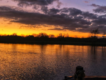 A strike orange band lights up the middle of the image, and is reflected in the river below. Above, the sky is indigo and dark clouds line the sky. Dark silhouettes of shrubs that line the water sit in the bottom third of the image