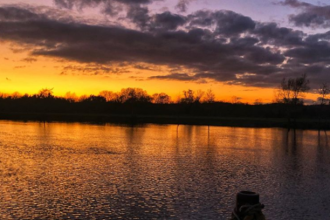 A strike orange band lights up the middle of the image, and is reflected in the river below. Above, the sky is indigo and dark clouds line the sky. Dark silhouettes of shrubs that line the water sit in the bottom third of the image