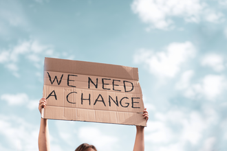 A person holds up a sign made out of cardboard which reads: "WE NEED A CHANGE."