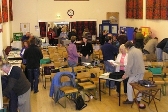 People at a conference in a town hall are stood at the edges of the room looking at artifacts and papers