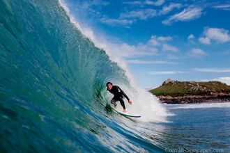 A male surfer in a black wetsuit, faces the camera and rides on his surfboard within a curling wall of a cresting wave, topped with white surf, in a sea coloured blue and turquoise.