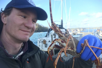 A man in a blue cap holds a crab in one blue gloved hand and smiles down at it