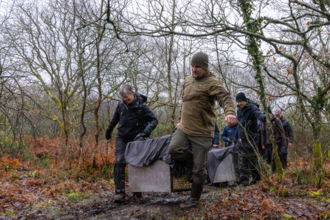 A group of people carry large covered crates through a muddy woodland area, transporting beavers to a release site among bare winter trees