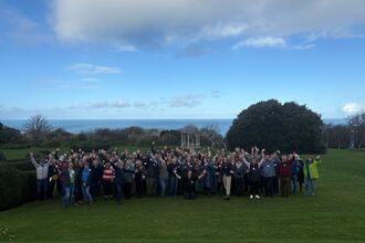  A group of people wave at the camera. They are stood on grass with the sea and some greenery in the background behind them. The sky is blue with some wispy clouds about.