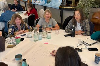 People sat around a round table at a conference chatting. One person is writing