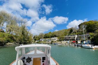 View from a small boat entering a calm river harbour, with moored sailboats, waterside houses, wooded hills, and a blue sky with scattered clouds.