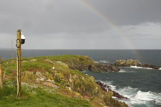 A rocky coastline overlooking the sea, with grey clouds in the background and a rainbow over the cliffs