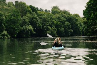 Two people in a kayak, paddling down a river surrounded by green trees