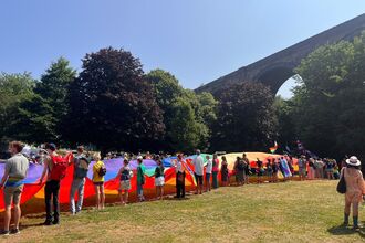 Large group of people standing on a grassy field beneath a tall stone viaduct on a sunny day, holding a long colourful Pride banner.