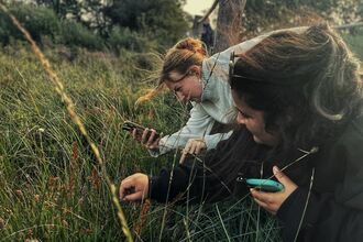 Two women are looking at some grass close up with phones in their hands.