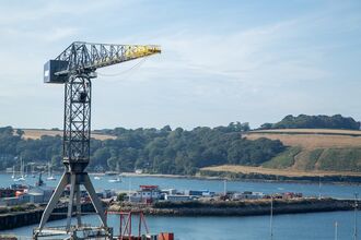 Port crane overlooking Falmouth Docks, with water, boats, and rolling green hills in the background.