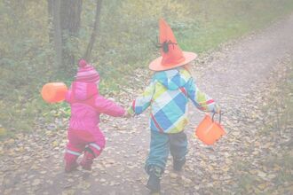 Two children run along a woodland path, facing away from the camera. They are wrapped up in winter clothing with orange buckets and the taller child on the right is wearing an orange pointy hat with a spider on. 