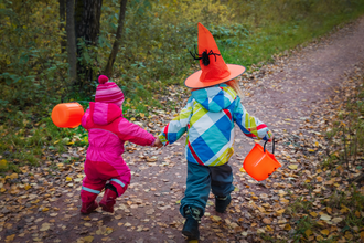 Two children run along a woodland path, facing away from the camera. They are wrapped up in winter clothing with orange buckets and the taller child on the right is wearing an orange pointy hat with a spider on. 
