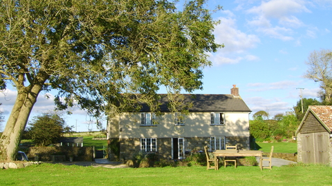 A sunny photograph of a detached farmhouse. A lawn is seen in the foreground, with a large tree to the house's left.