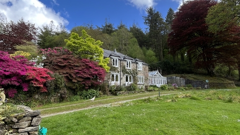 A sunny shot of The Lodge. A farmhouse can be seen in the background, surrounded by an array of brightly-coloured trees and shrubs.