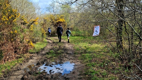A family in the distance walking through a muddy footpath. A sign in the bottom left says 'this way' with an arrow pointing to where the family are walking