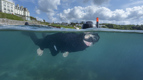 A man wearing a wetsuit is visible snorkelling underwater, with buildings visible on the horizon behind him