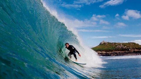 A male surfer in a black wetsuit, faces the camera and rides on his surfboard within a curling wall of a cresting wave, topped with white surf, in a sea coloured blue and turquoise.