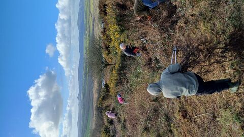 Group of people clearing vegetation on a hillside with scenic countryside and hills in the background under a blue sky with scattered clouds.