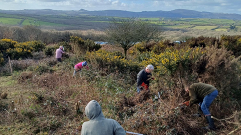 Group of people clearing vegetation on a hillside with scenic countryside and hills in the background under a blue sky with scattered clouds.