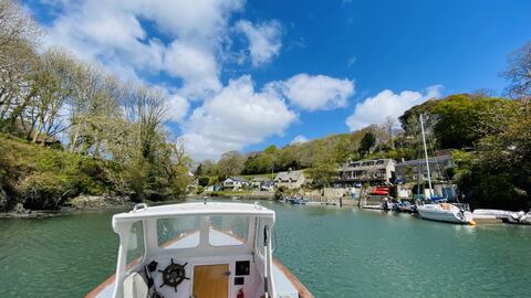 View from a small boat entering a calm river harbour, with moored sailboats, waterside houses, wooded hills, and a blue sky with scattered clouds.