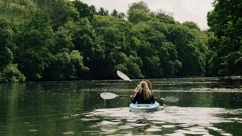 Two people in a kayak, paddling down a river surrounded by green trees
