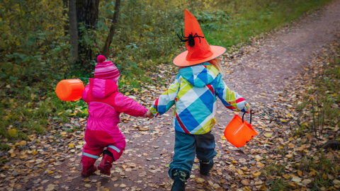 Two children run along a woodland path, facing away from the camera. They are wrapped up in winter clothing with orange buckets and the taller child on the right is wearing an orange pointy hat with a spider on. 