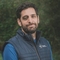 Man with dark hair and beard wearing a navy blue puffer vest over a blue shirt, standing outdoors in front of a blurred green background