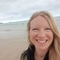 A blonde haired woman smiles at the camera with the sea in the background 