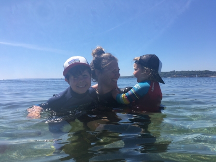 Marine Team Officer Abby Crosby bobs in the water with her two young children, surrounded by clear,calm blue waters and a bright blue sky behind. They are all laughing happily and enjoying the sun and the sea.