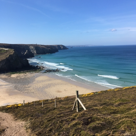 a picture-perfect postcard image, the coast path trails off in the left hand corner, the green heather rolls down to a sandy yellow beach where the tide is out and aquamarine waves roll into the shore. The aquamarine blue gradually darkens and meets a light blue sky above