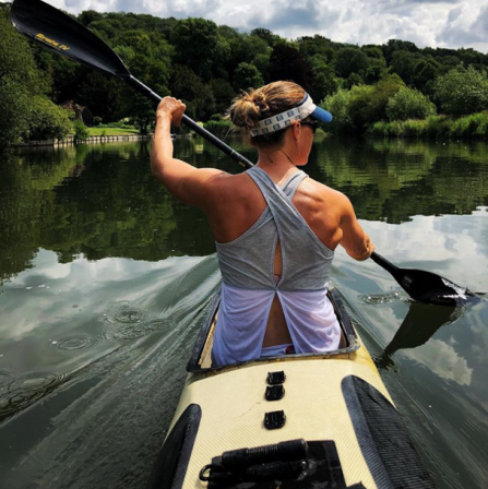 Helen sits in a canoe with her back to the camera, paddling through a calm river with lush vegetation in the hills beyond