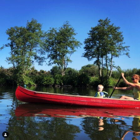 A bright blue sky reflects in the river, and three tall trees stretch up in the background. The sky and river are divided by a bright red canoe which Helen is paddling in, as her son looks out towards the camera