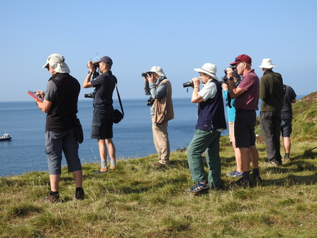 A group of people stood on a grassy headland looking out to sea with binoculars and clipboards