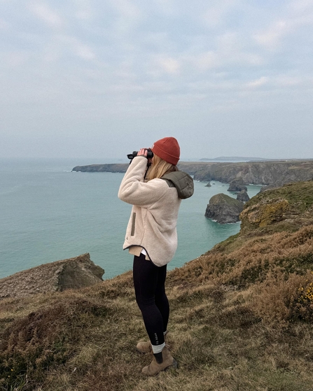 A woman stands on the clifftop with binoculars in hand, looking out to sea