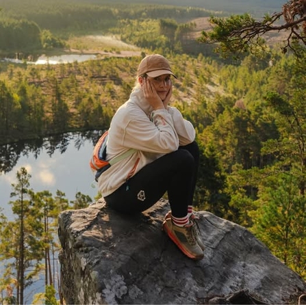 A woman is sat on a rock with an expansive view of pine trees and water below here. She is wearing a white jumper, black leggings and a cap, she cups her face in her hands