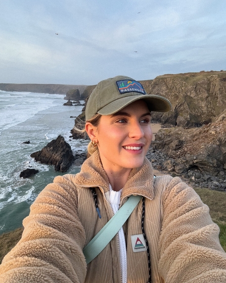 A woman smiles taking a selfie, eyes looking to the left, with cliffs and waves of Cornwall's coast in the background