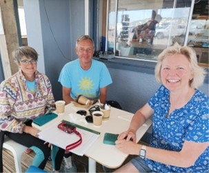 Three people sit at an outdoor table with coffee cups, a camera, and green objects. They are smiling and casually dressed.