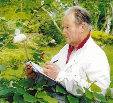 A man stands surrounded by greenery drawing a flower. He is wearing a white jacket with a red shirt underneath. He holds the pen and draws with his left hand.