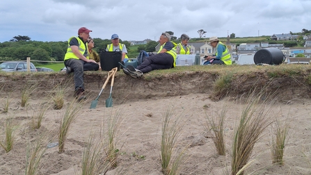 Volunteers sat on the edge of a small sand dune, some marram grass visible in the foreground. The people are wearing hi-vis jackets