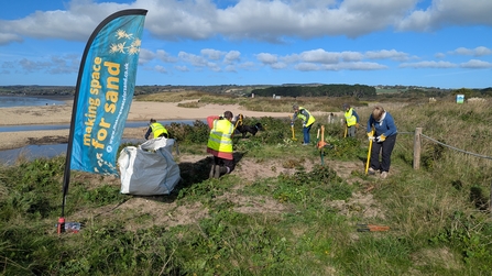 A flag saying 'making space for sand' waves on the dunes atop a beach, volunteers in hi-vis in the background are digging plants