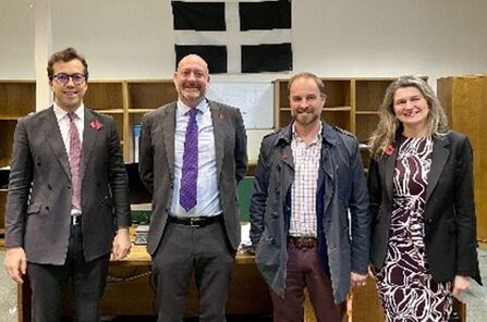 Four people standing indoors in front of a desk and wooden shelving units. A black-and-white flag is displayed on the wall behind them. The individuals are dressed in formal and semi-formal attire, including suits, ties, and patterned dresses. The setting appears to be an office or meeting room.