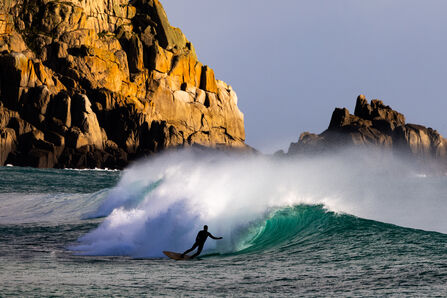 A surfer rides a wave under a rocky cliff face which is in the sun
