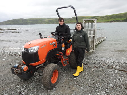 A man and woman on a small orange tractor with a long trailer on the back, on the shore