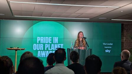 Alison Smith stands behind a podium with a green backgound behind, displaying the pride in our planet awards logo