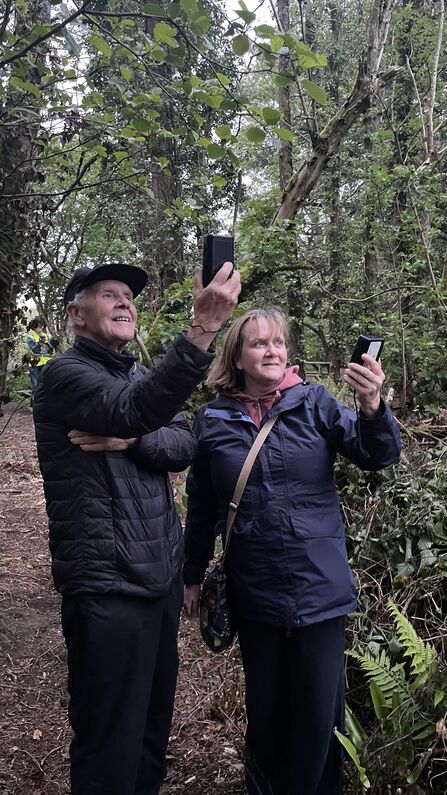 Two people in coats in a woodland holding small black devices up towards the trees