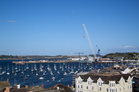 View across Falmouth Harbour showing numerous moored boats, a dockside crane, and waterside buildings under a clear blue sky.