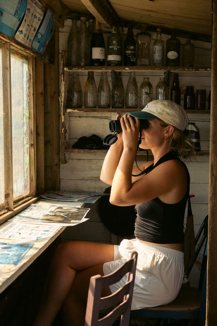 A woman sits in a ramshackle shed, looking out of the windows with binoculars