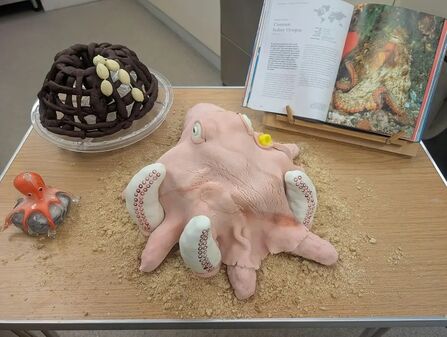 An octopus shaped cake on a table with an open book and a pot-shaped cake